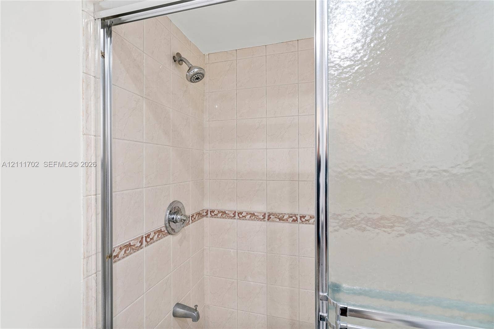Shower stall featuring square wall tile, decorative inset tile trim, chrome shower head, chrome single-handle mixing valve, and a metal-framed glass sliding door