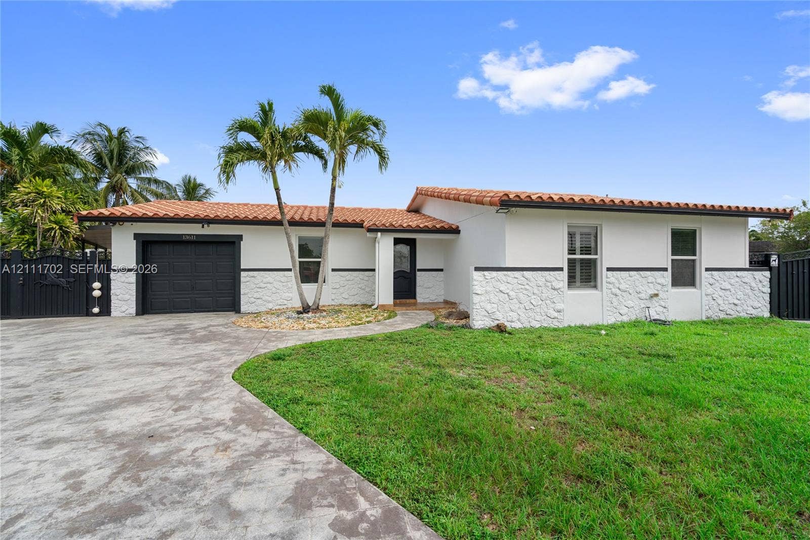 Single-story exterior featuring white stucco walls, textured stone wainscoting, a terracotta tile roof, a single-car garage, and mature palm trees