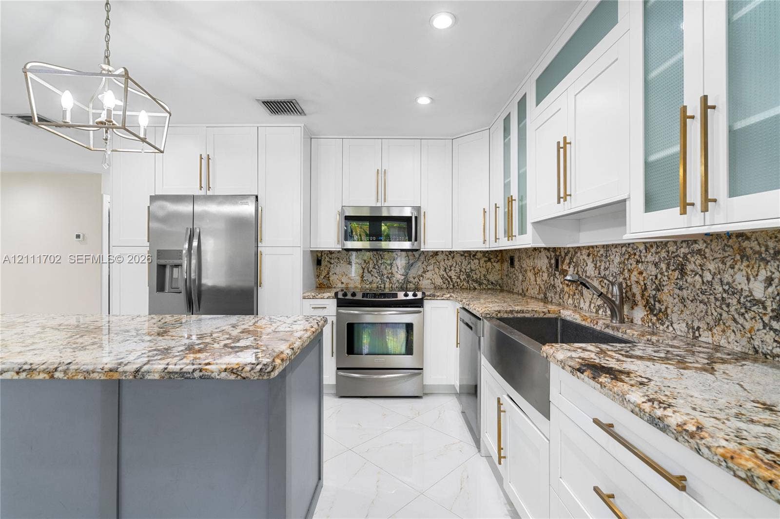 Kitchen featuring white shaker cabinetry, stone countertops, stainless steel farmhouse sink, brushed gold hardware, and a center island with contrasting gray cabinetry