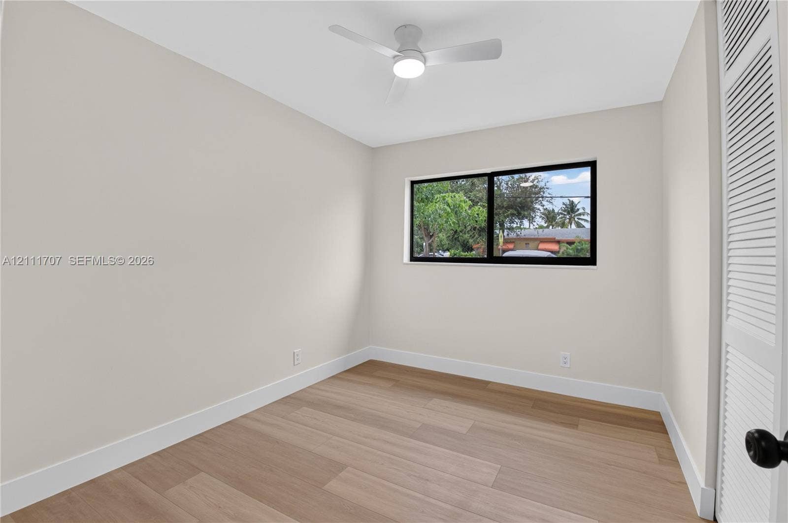 301 SW 12th Avenue, Delray Beach, FL 33444 Neutral-toned interior featuring wood-finish flooring, a white ceiling fan with integrated lighting, a sliding window with a black frame, and a louvered bi-fold closet door