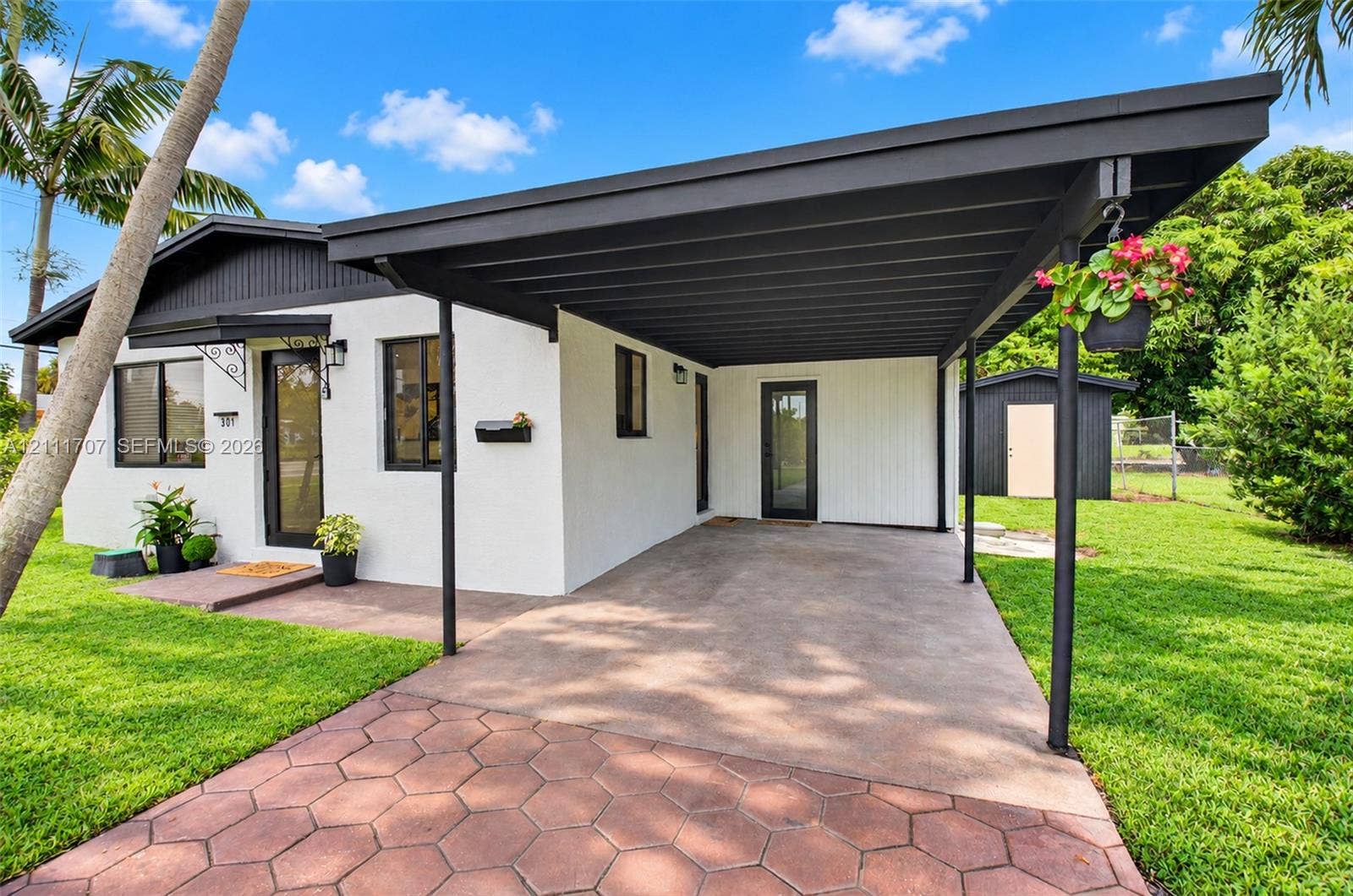 301 SW 12th Avenue, Delray Beach, FL 33444 Exterior featuring a white stucco facade, covered carport with dark-toned trim, hexagonal paver driveway, and detached storage shed