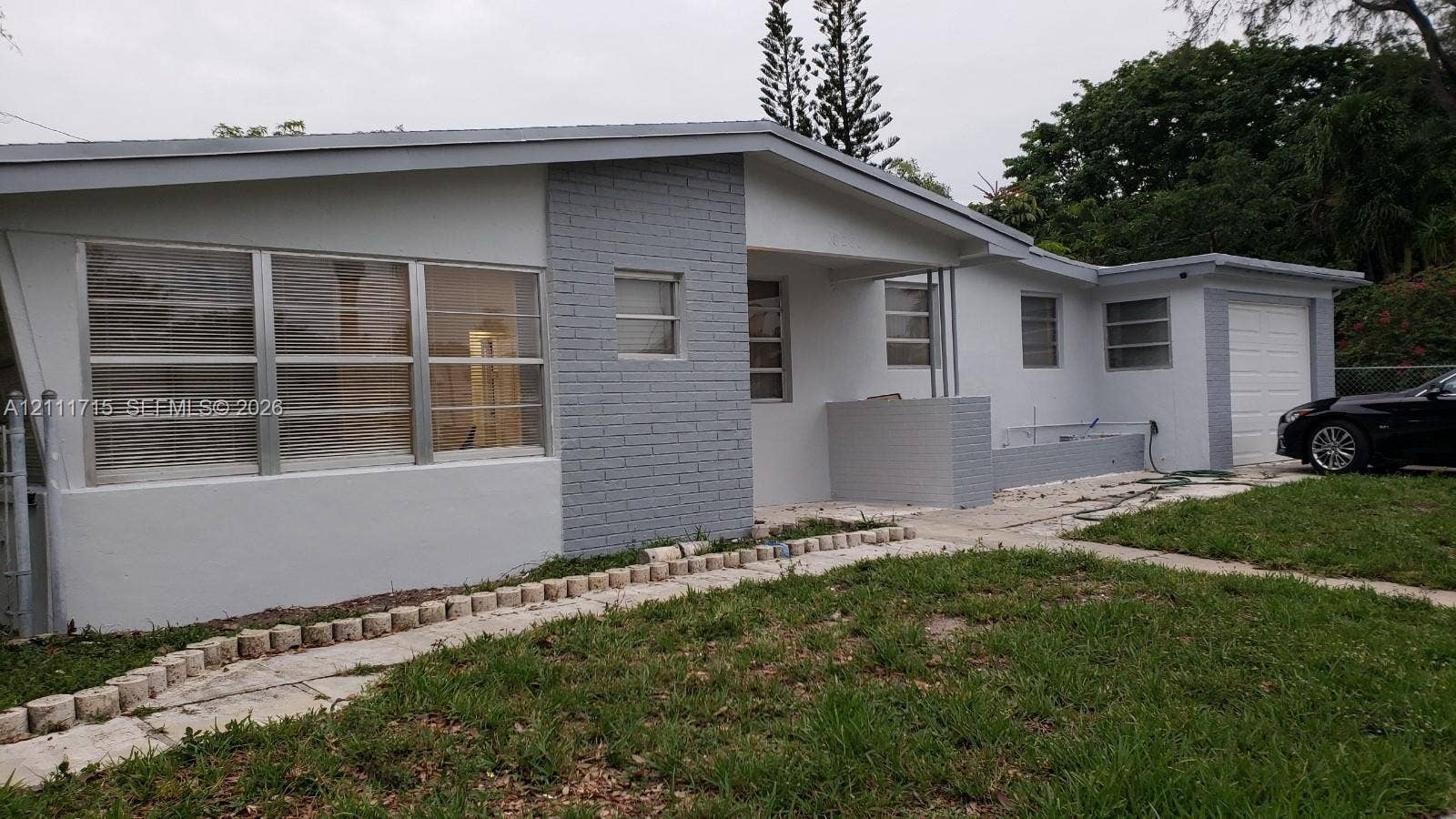18280 NE Glades Drive, North Miami Beach, North Miami Beach, FL 33162 Single-story exterior featuring painted brick accents, multi-pane horizontal windows, an attached garage, and a stone-bordered concrete walkway