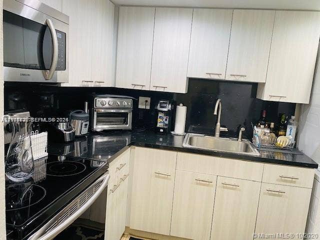 Kitchen featuring light-toned cabinetry with linear hardware, black solid-surface countertops, black backsplash, stainless steel sink with a goose-neck faucet, and stainless steel appliances