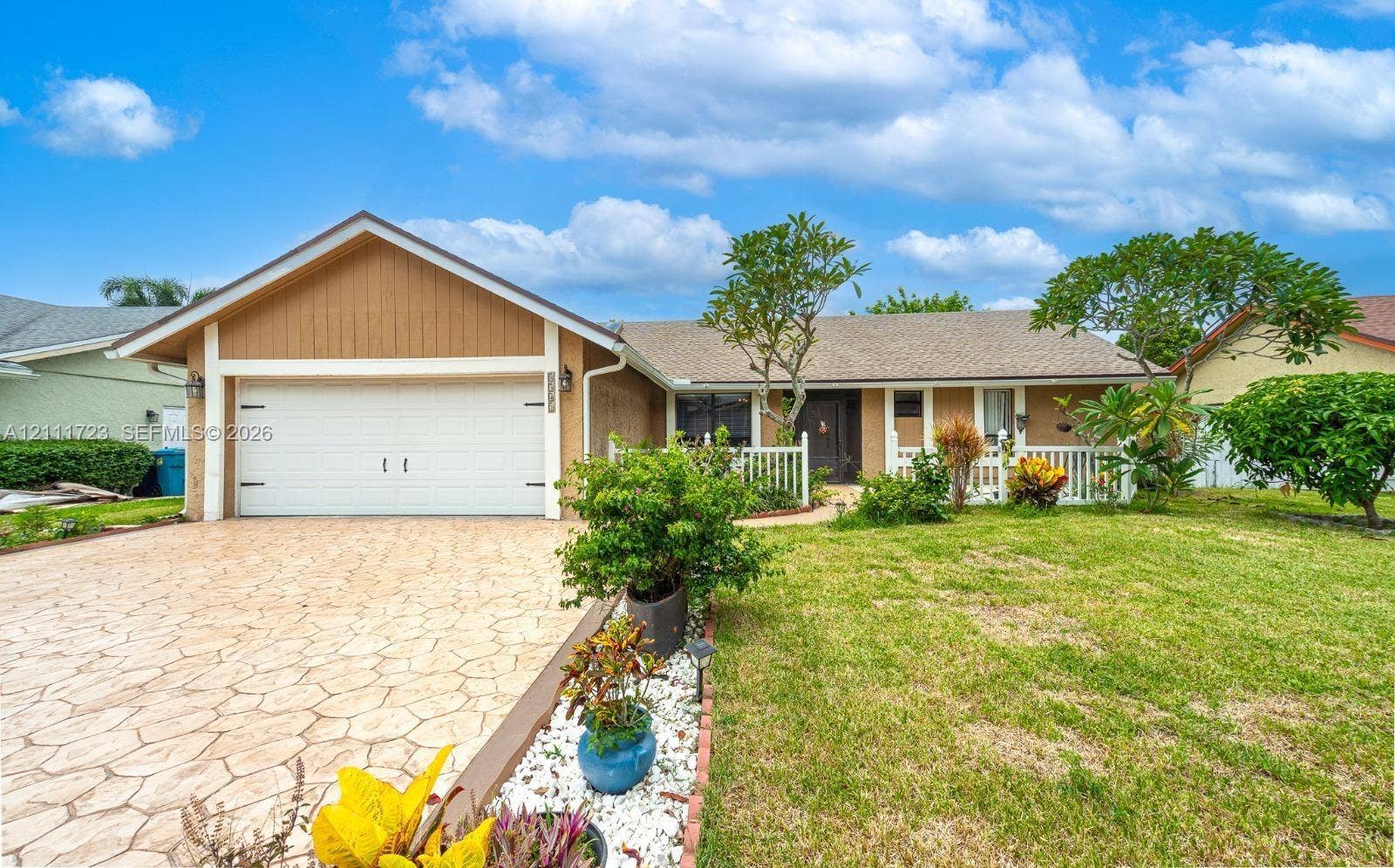 8541 NW 51st Court, Lauderhill, FL 33351 Single-story exterior featuring a stamped concrete driveway, two-car garage, white picket fencing, and mature landscaping
