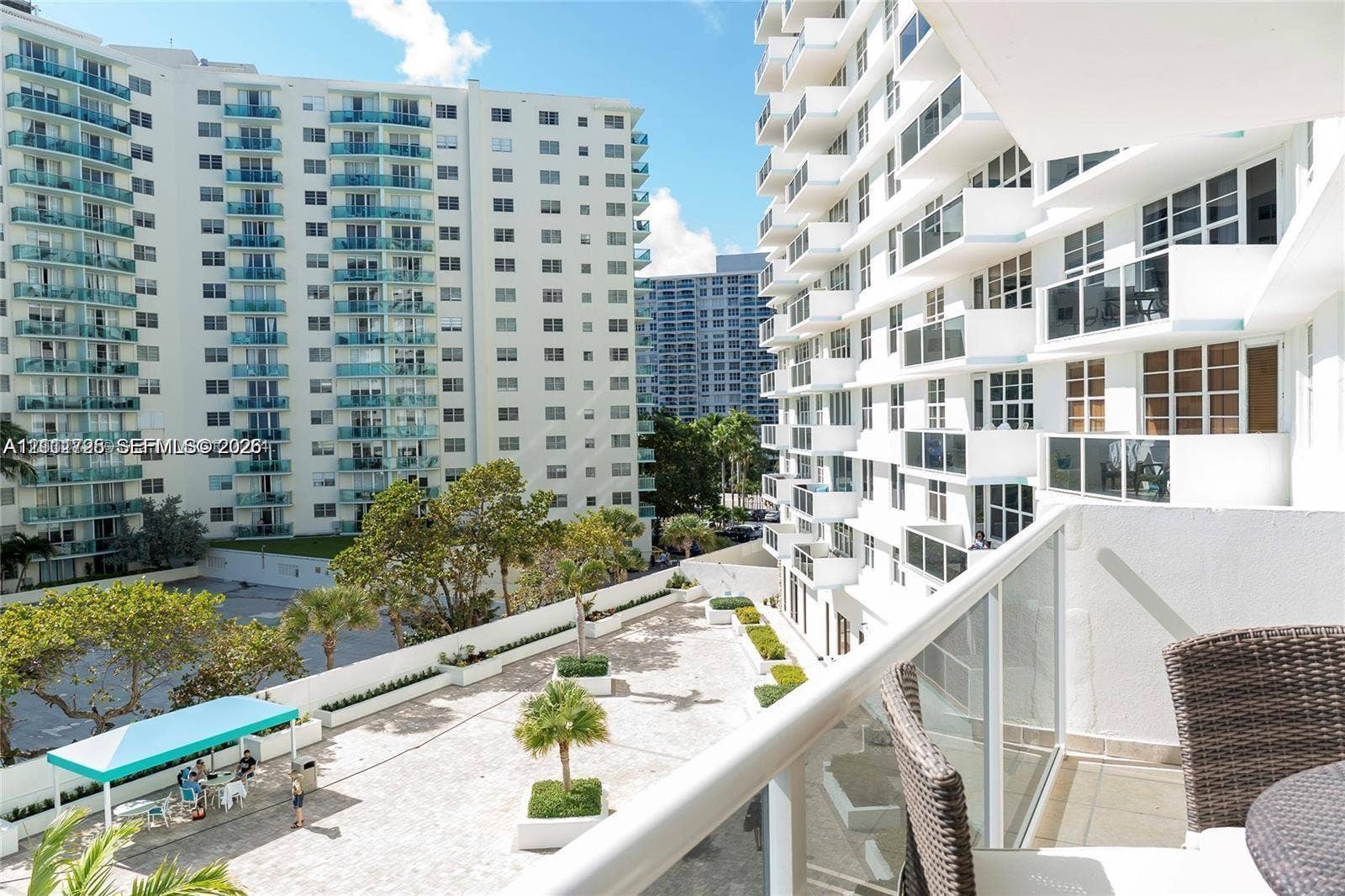 3725 S Ocean Drive, #508, Hollywood, Hollywood, FL 33019 Private balcony with glass panel railing overlooking a paved outdoor terrace featuring mature palms, raised planters, and a shaded seating area