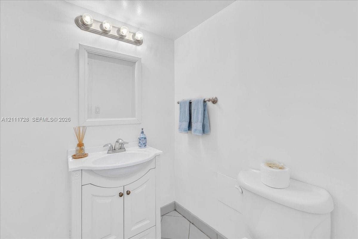 White vanity with a molded countertop and integrated basin, framed wall mirror, four-light vanity fixture, and neutral tile flooring