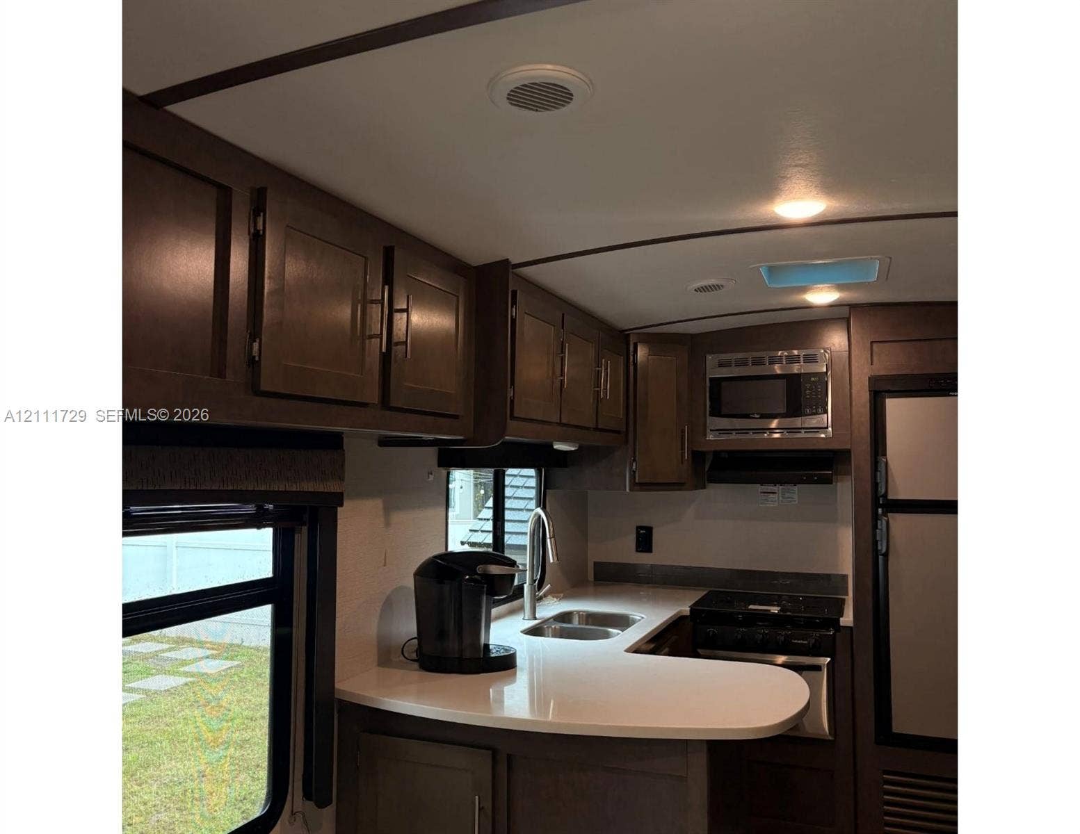 Kitchen featuring dark wood cabinetry, light-toned solid surface countertops, stainless steel dual-basin sink with gooseneck faucet, built-in microwave, and stainless steel refrigerator