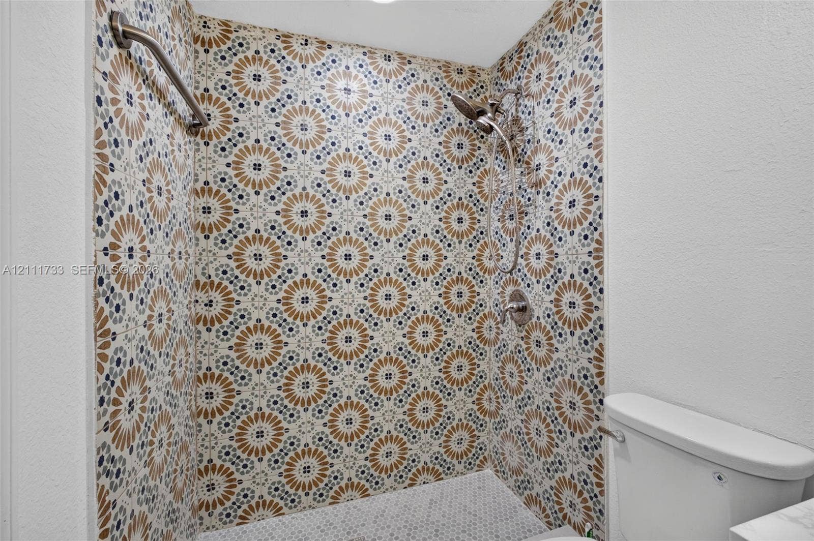 Walk-in shower featuring decorative patterned tile walls, a handheld showerhead, a metal grab bar, and a mosaic tile floor