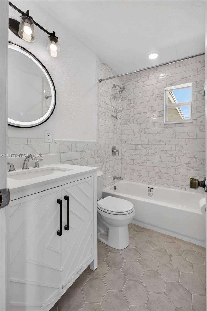 Main Bathroom featuring a white chevron-pattern vanity with a solid-surface countertop, an illuminated round mirror, porcelain tile shower surround, and hexagonal floor tile