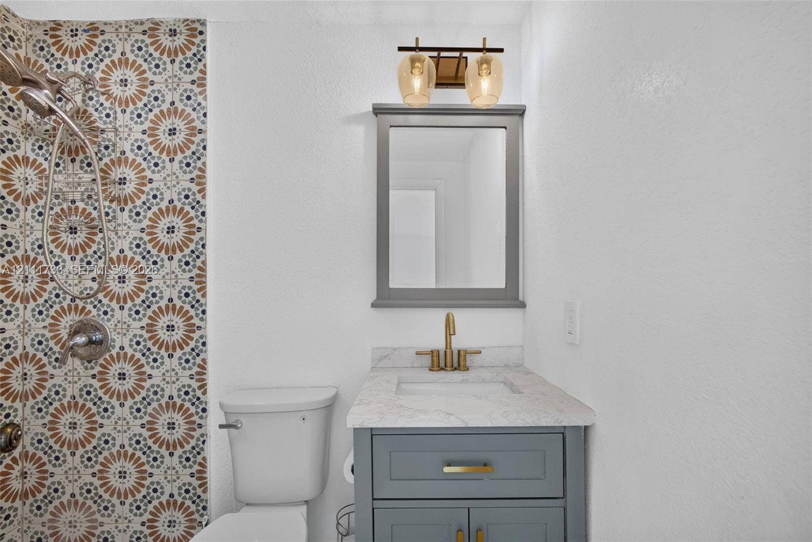 Ensuite Bathroom featuring patterned tile shower surround, grey vanity countertop, undermount sink, metallic faucet, and a wall-mounted light fixture.