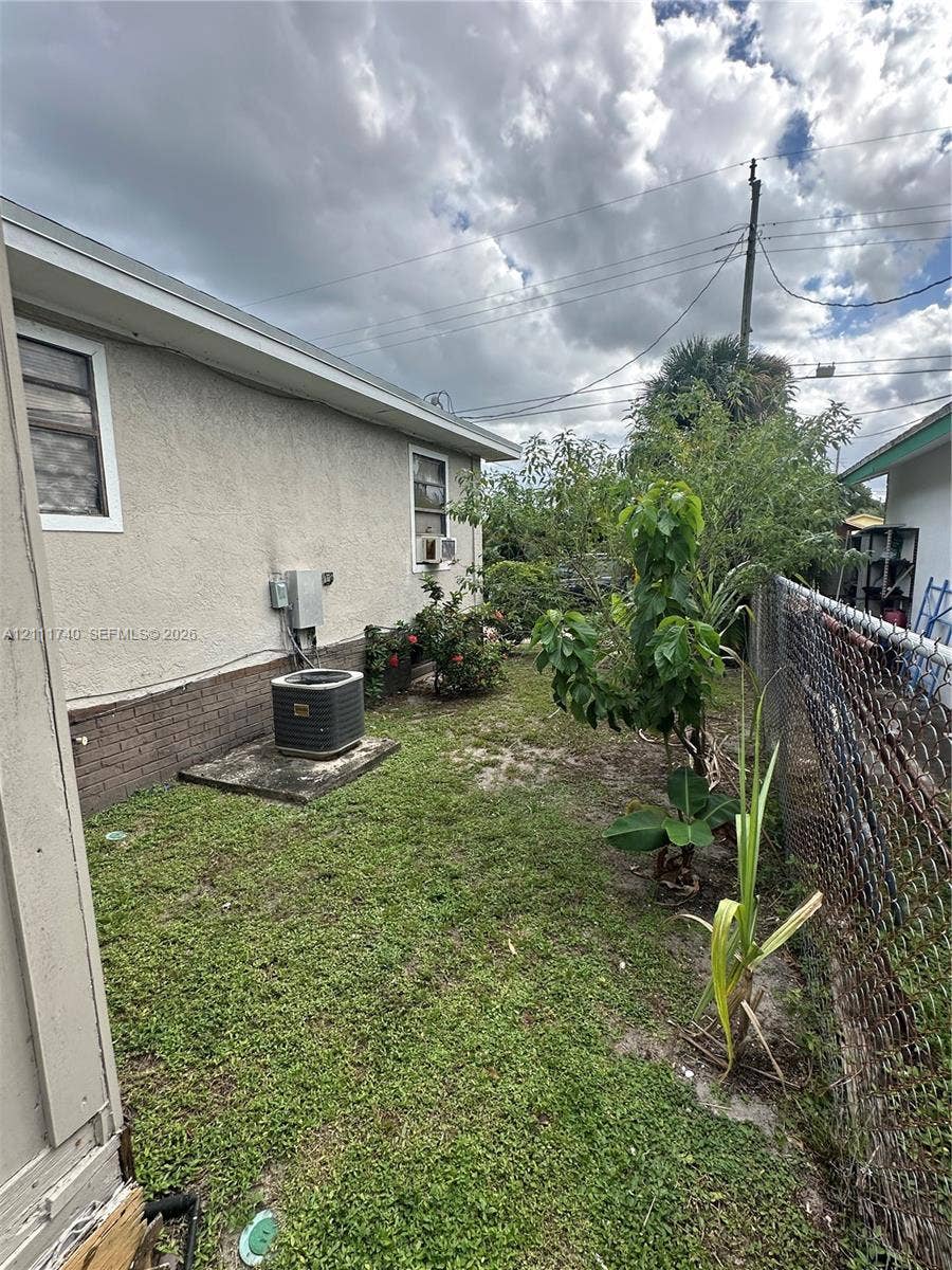 Exterior side yard featuring a stucco and brick facade, central air conditioning unit on a concrete pad, and a chain-link perimeter fence