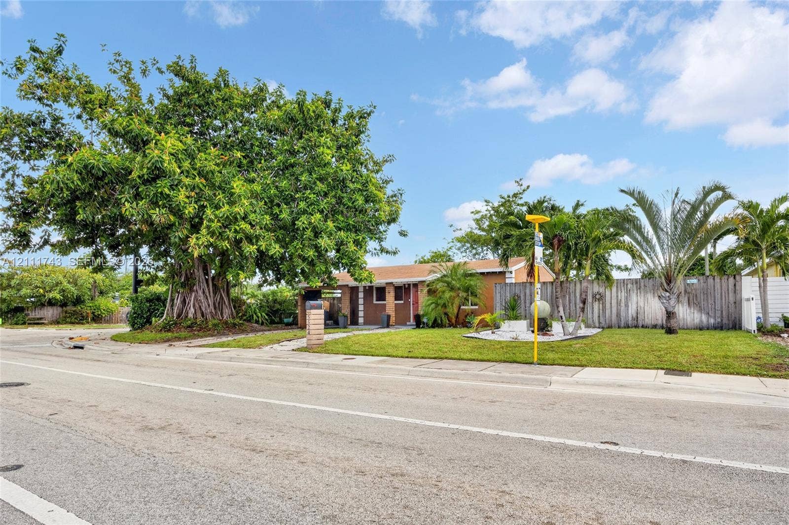 5524 N Andrews Avenue, Oakland Park, FL 33309 Single-story residence featuring a horizontal wood-slat privacy fence, manicured front lawn, mature specimen tree, and tropical landscaping