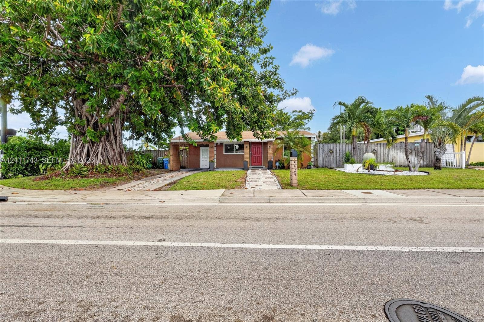 5524 N Andrews Avenue, Oakland Park, FL 33309 Single-story residence featuring a masonry exterior, red entry door, mature canopy tree, manicured lawn, and wood-style privacy fencing