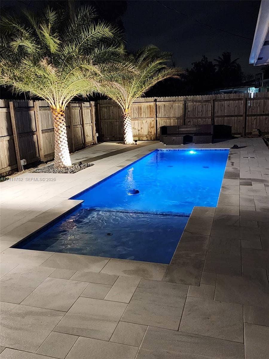 2901 NE 11th Terrace, Pompano Beach, Pompano Beach, FL 33064 Rectangular pool featuring a shallow entry shelf, expansive stone paver decking, mature palm trees, and perimeter wood privacy fencing