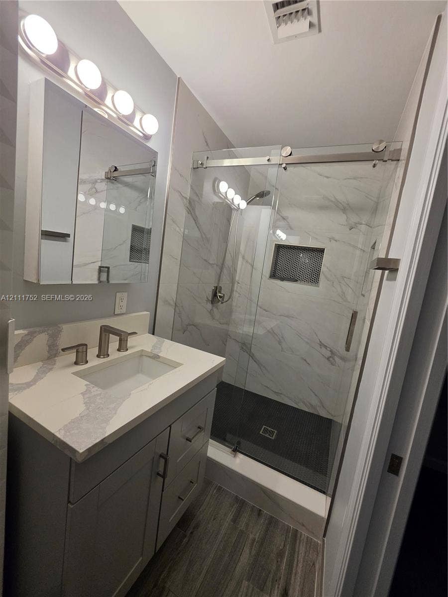 2901 NE 11th Terrace, Pompano Beach, Pompano Beach, FL 33064 Bathroom vanity featuring a light-toned countertop, rectangular undermount sink, and brushed-finish fixtures, paired with a glass-enclosed walk-in shower with stone-patterned wall tile, a mosaic-tiled recessed niche, and wood-finish flooring
