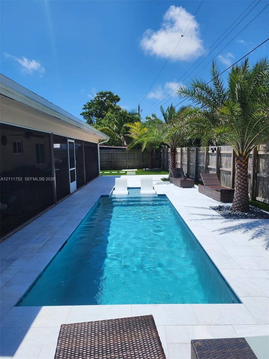 2901 NE 11th Terrace, Pompano Beach, Pompano Beach, FL 33064 Rectangular swimming pool featuring a Baja shelf, light-toned stone pool deck, and perimeter wood fencing accented by mature palm trees
