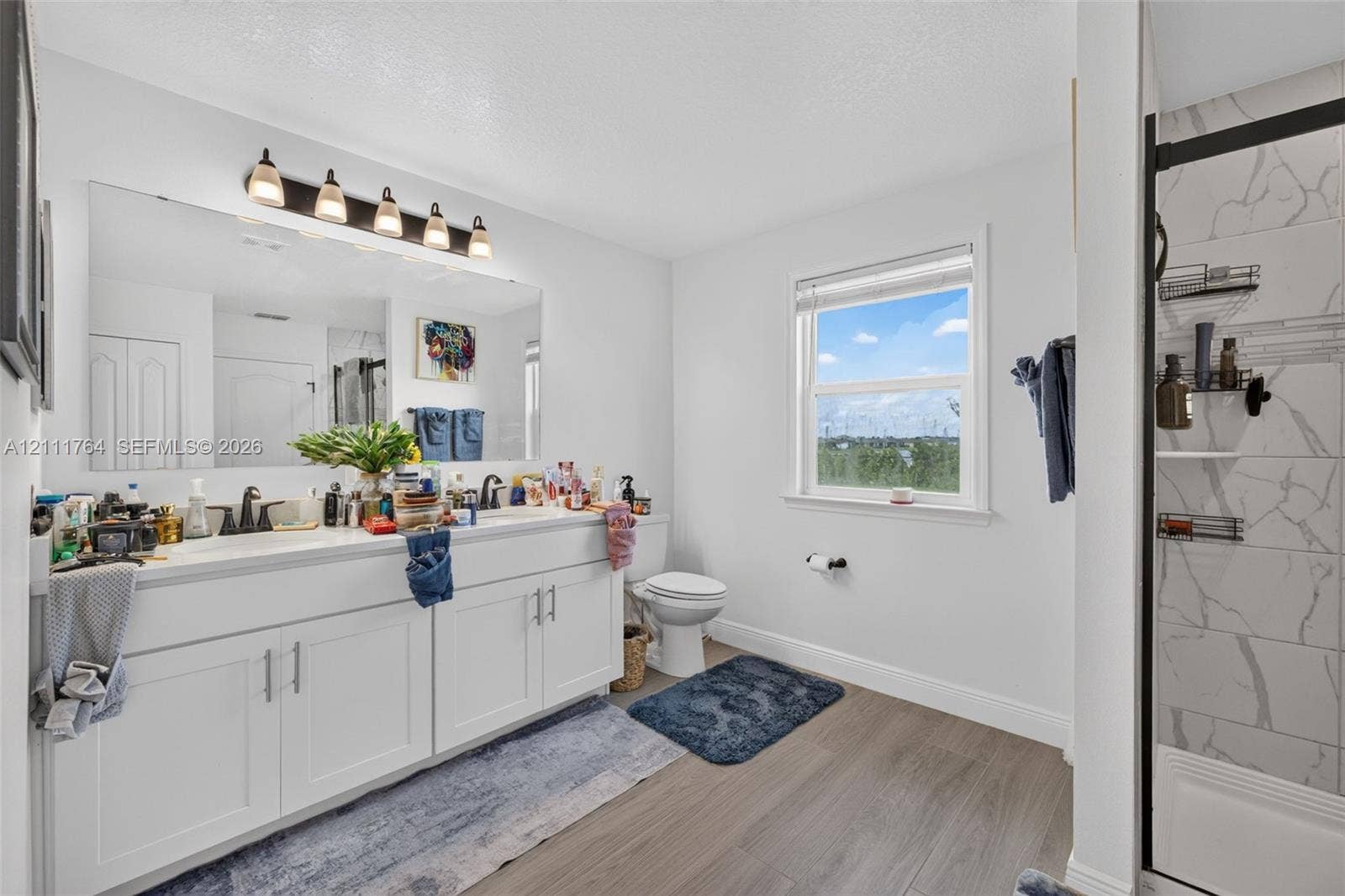 Bathroom featuring a dual-sink vanity with white cabinetry, a full-width wall mirror with five-bulb overhead lighting, wood-finish flooring, and a walk-in shower with stone-patterned tile surround and a black-frame glass door