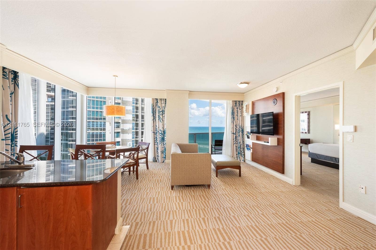Open-concept living space featuring floor-to-ceiling windows, built-in wooden media cabinetry, a kitchen island with stone countertop, and a private balcony with water scenery