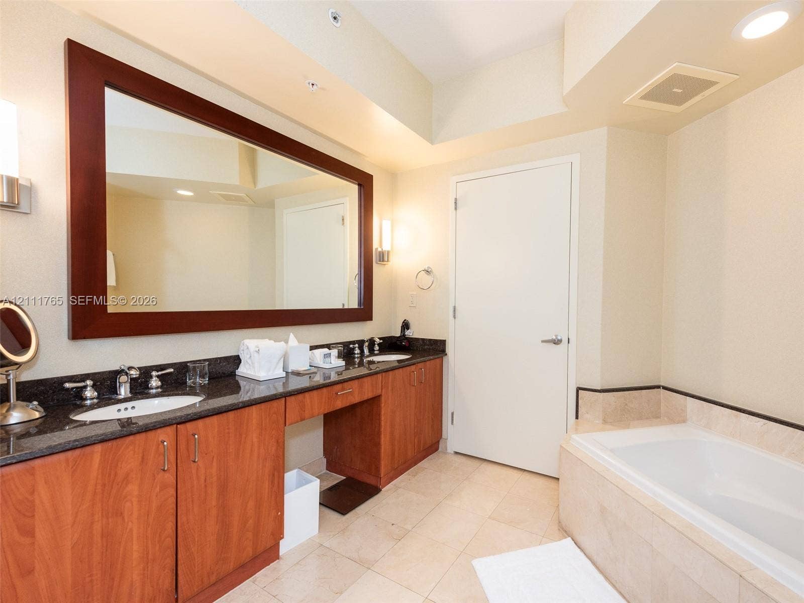 Dual-sink vanity featuring wood cabinetry, stone countertops, and a large wall-mounted mirror, complemented by a tiled surround soaking tub