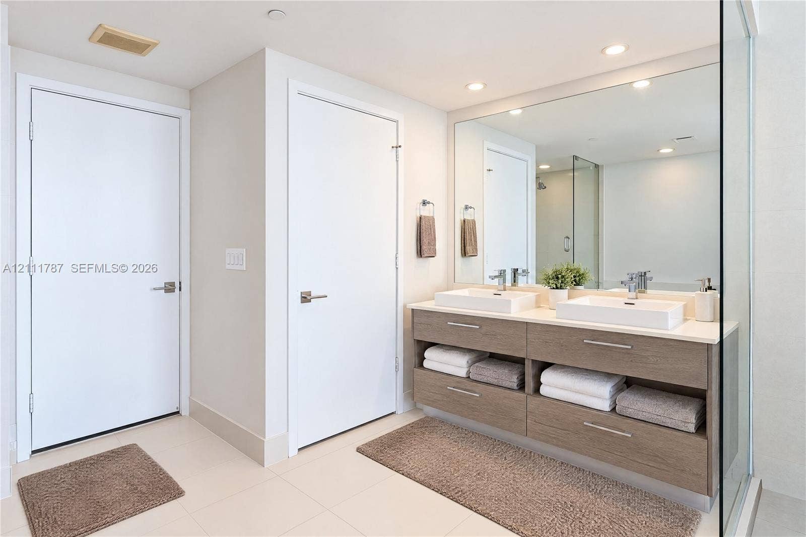 Dual-sink vanity with wood-finish cabinetry, rectangular vessel sinks, light-toned stone countertop, large wall-to-wall mirror, and glass-enclosed shower
