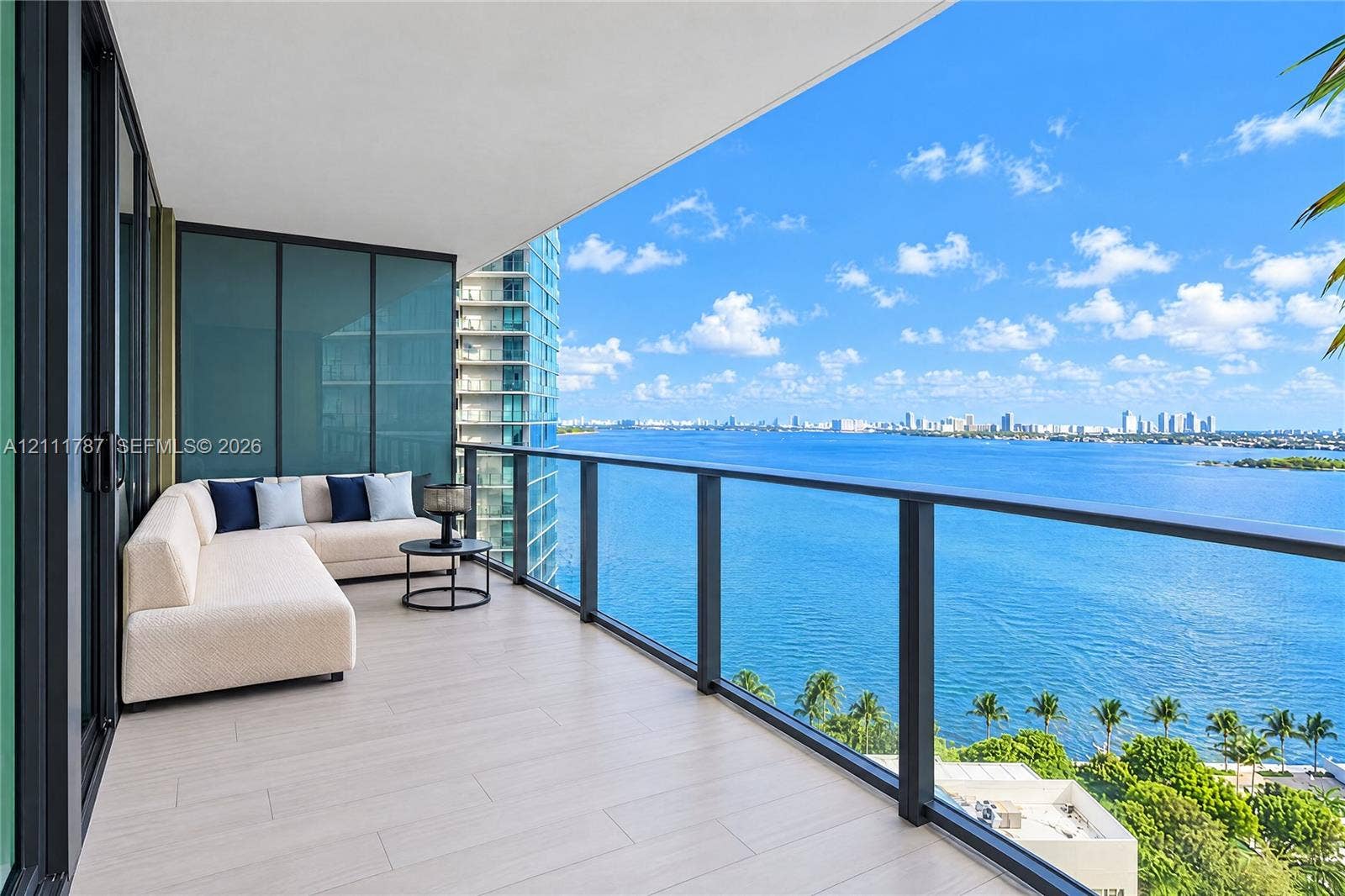 Covered balcony featuring wood-finish tile flooring, matte black metal railings, and floor-to-ceiling glass paneling with panoramic water and skyline scenery