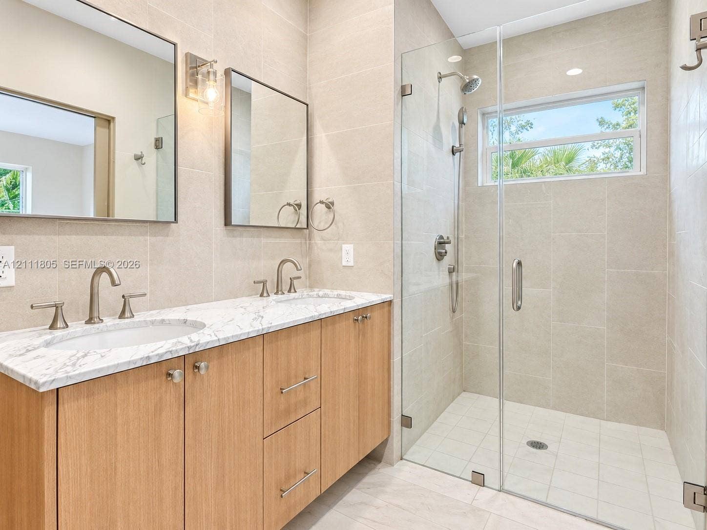 Double vanity featuring natural wood-finish cabinetry, a stone-patterned countertop, dual under-mount sinks, frameless glass-enclosed walk-in shower, and large-format wall tiling