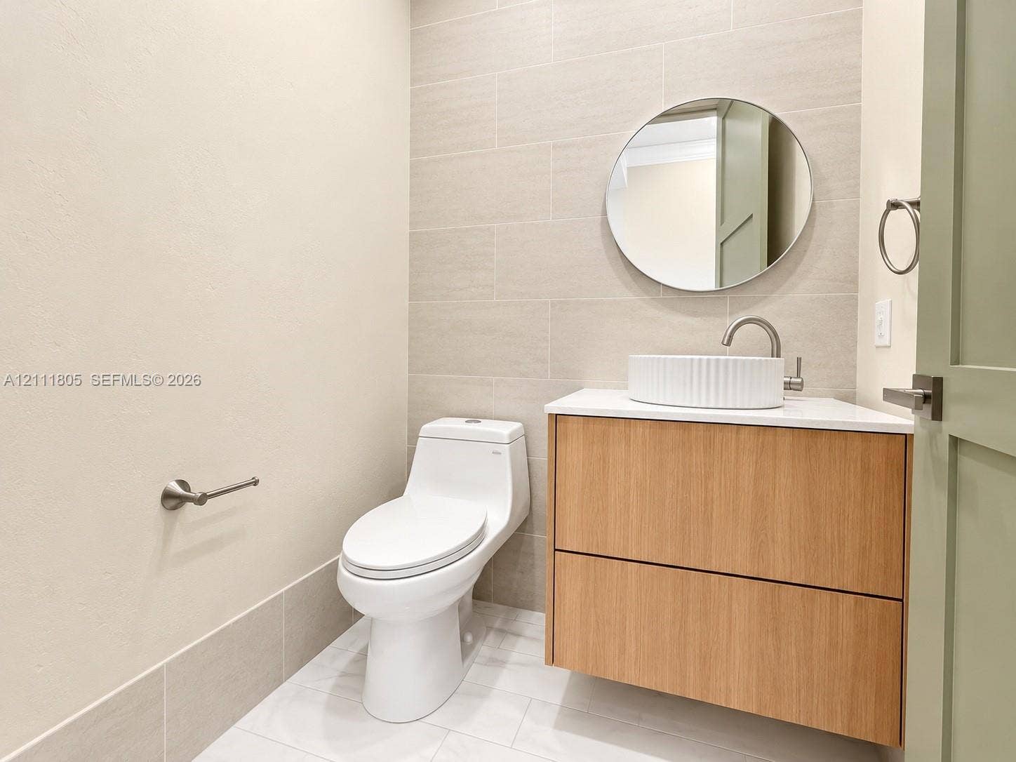 Floating wood-finish vanity with a white vessel sink, neutral rectangular wall tile, round mirror, and one-piece toilet
