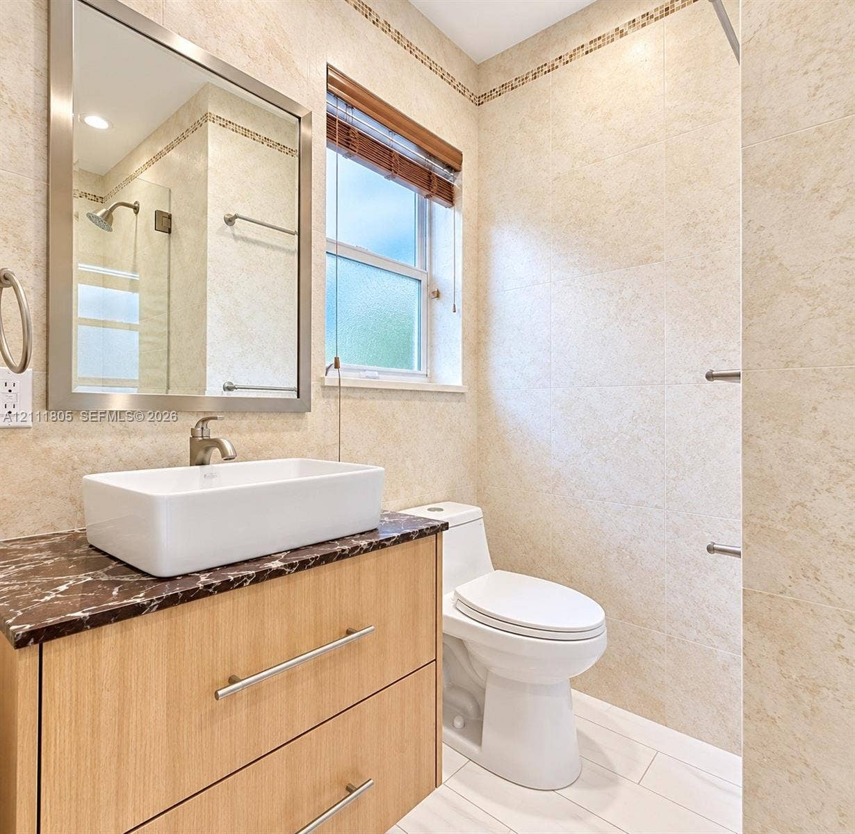 Bathroom featuring a vanity with a light wood-finish, stone countertop, rectangular vessel sink, wall-mounted mirror with a metallic frame, and full-height wall tiling with decorative border inlay