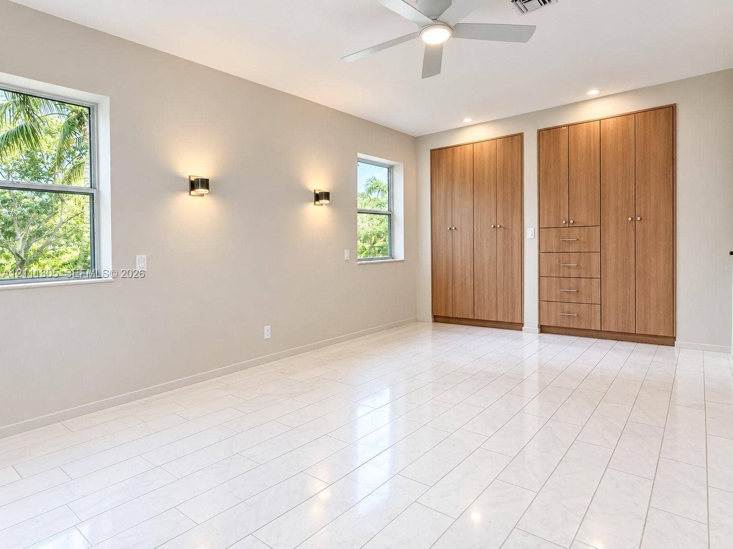 Neutral-toned interior featuring large-format tile flooring, built-in floor-to-ceiling wood-finish cabinetry, wall-mounted light fixtures, and a ceiling fan with integrated lighting
