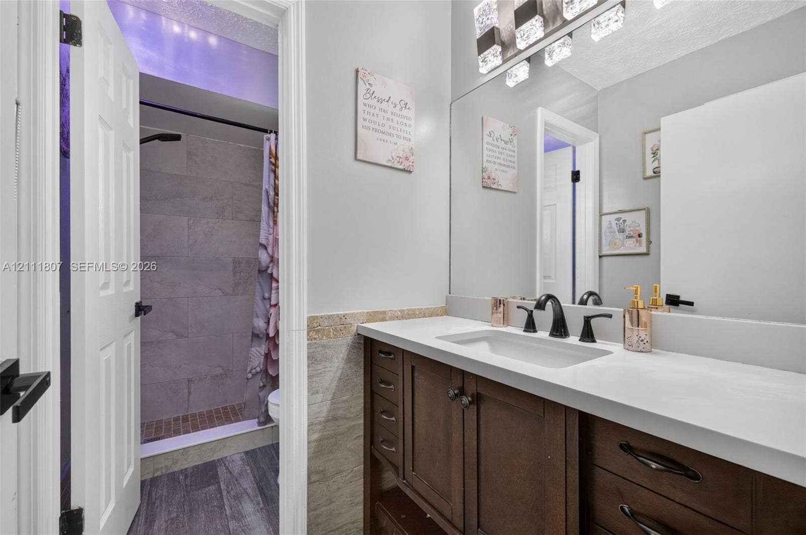 Bathroom featuring a dark wood vanity with white countertop, undermount sink, matte black widespread faucet, large wall-mounted mirror with integrated lighting, and a walk-in shower with grey tile surround