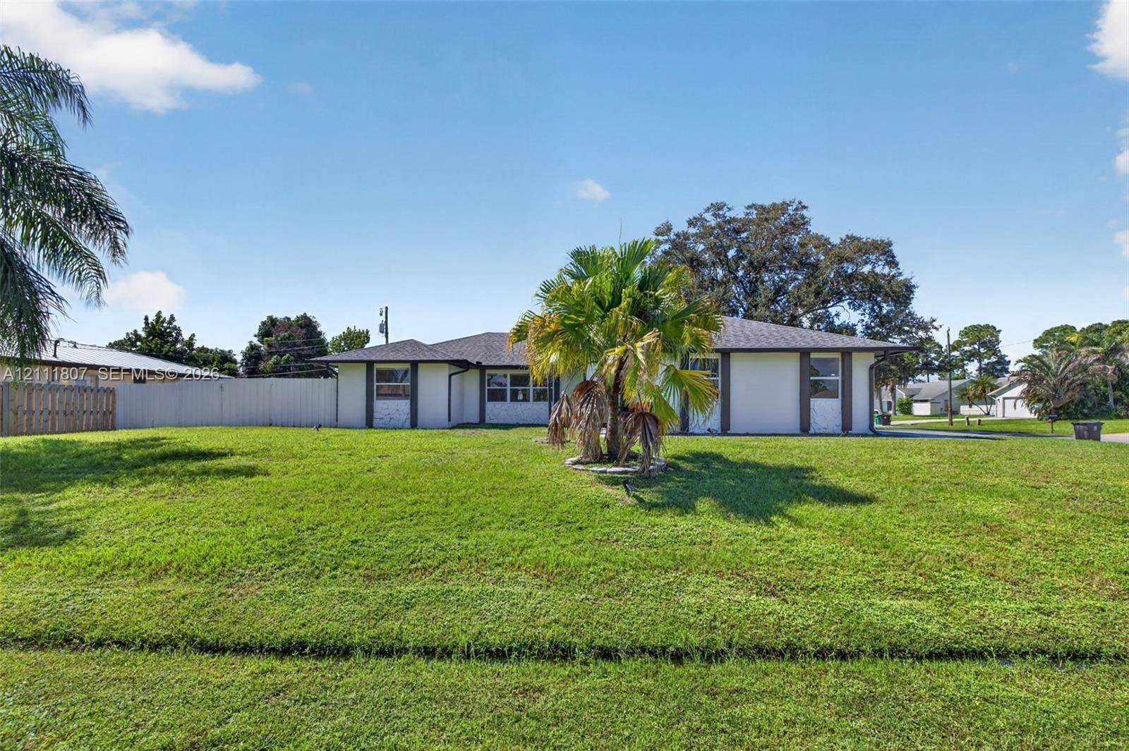 Single-story exterior featuring a light-toned stucco facade, shingled hip roof, expansive grassy lawn, and privacy fencing