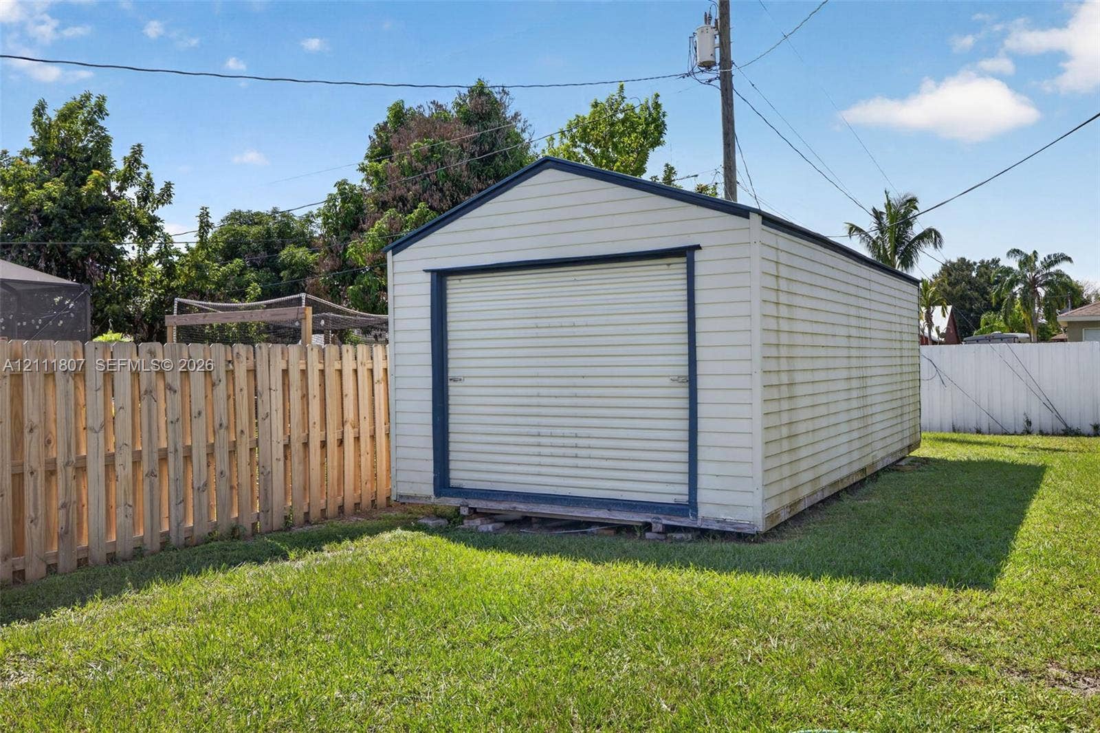 Detached storage structure with metal siding, a roll-up garage door, and a gabled roof, situated on a lawn enclosed by wood and vinyl privacy fencing