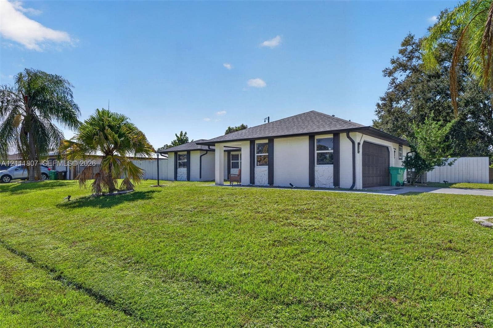 Single-story exterior featuring a light-colored stucco facade, contrasting dark trim accents, a multi-paneled garage door, a covered entryway, and an expansive lawn