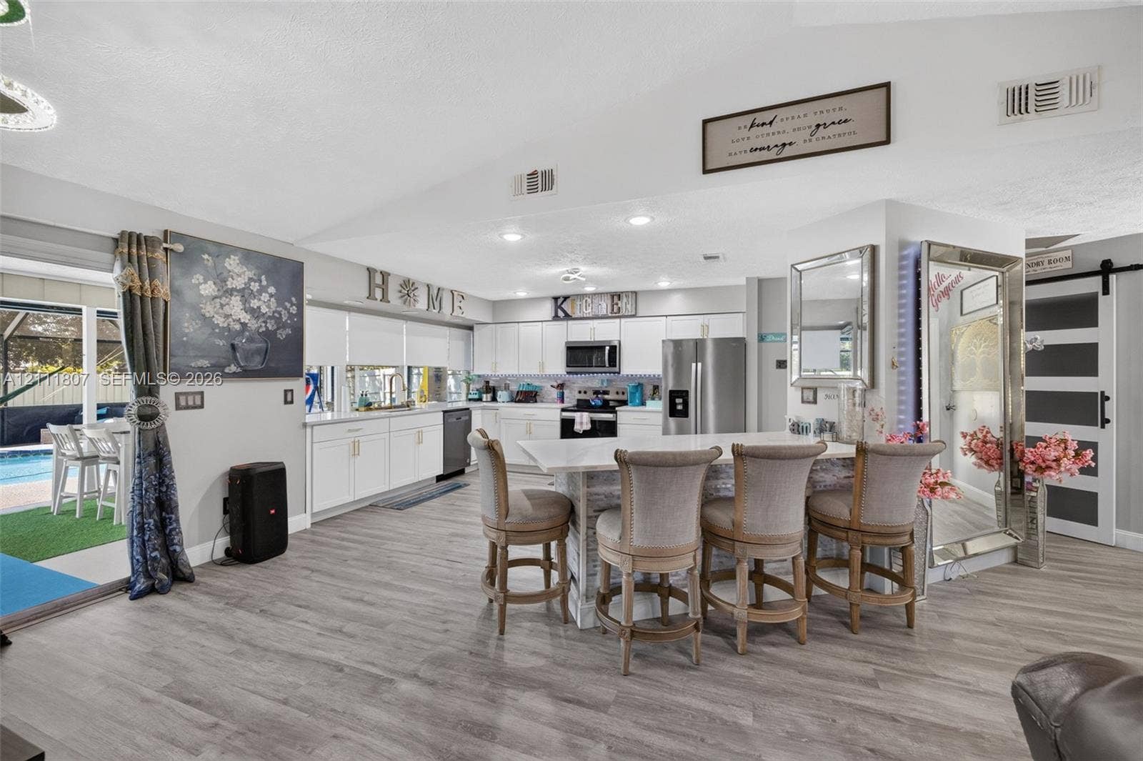 Open-concept kitchen featuring white shaker-style cabinetry, stainless steel appliances, stone-surface central island with bar seating, and wood-finish flooring