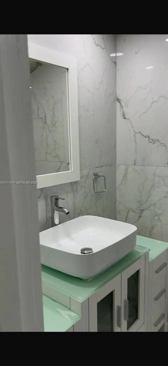 Bathroom featuring a vessel sink with a single-handle faucet, glass-top vanity with built-in cabinetry, framed mirror, and floor-to-ceiling veined tile wall surfacing