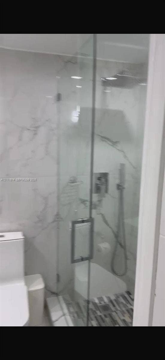 Walk-in glass-enclosed shower featuring large-format stone-patterned wall tile, rain-style shower head, handheld sprayer, and mosaic tile floor inlay