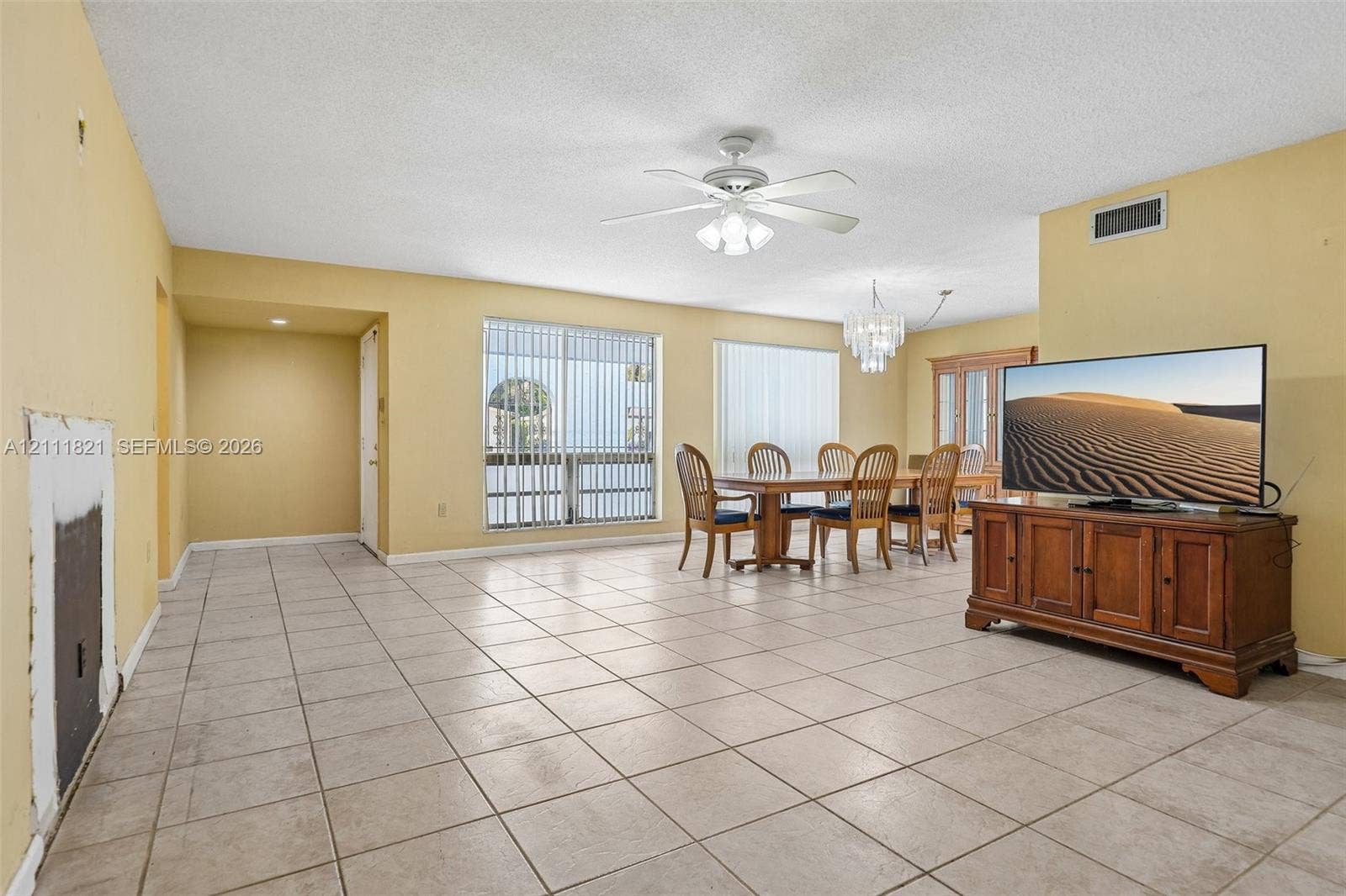 Neutral-toned tile flooring throughout the open-concept living area, featuring a decorative tiered chandelier, a white ceiling fan with integrated lighting, and large windows with vertical blinds