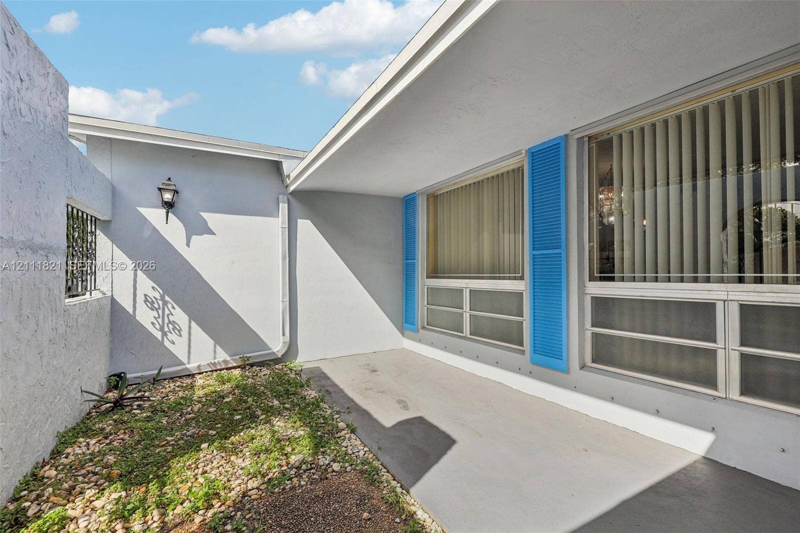 Covered exterior patio featuring concrete flooring, stucco siding, decorative blue shutters, and a wall-mounted carriage-style light fixture
