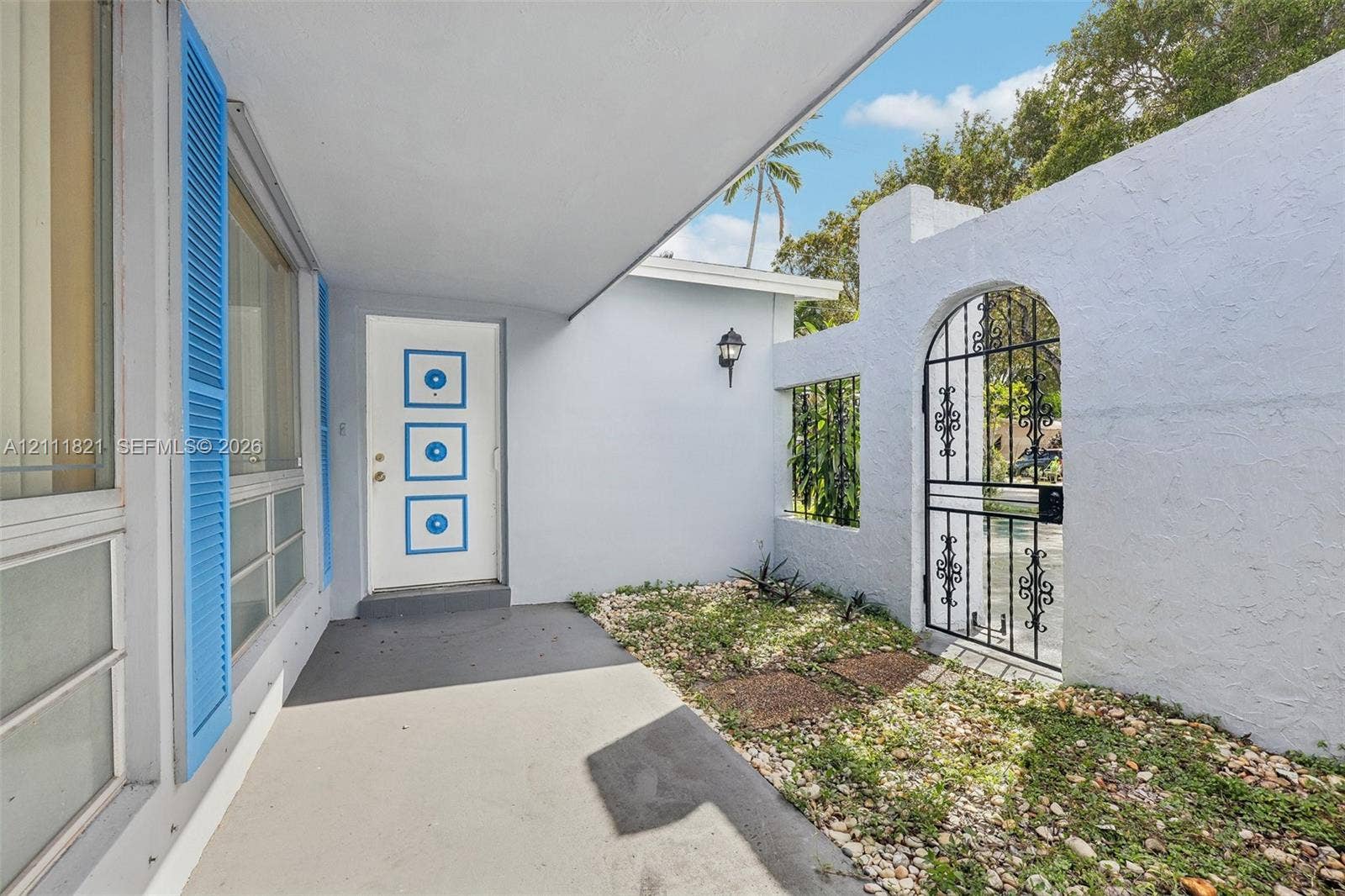 Covered entryway featuring a white door with geometric accent detailing, blue decorative shutters, and a gated courtyard entrance with an arched metal gate