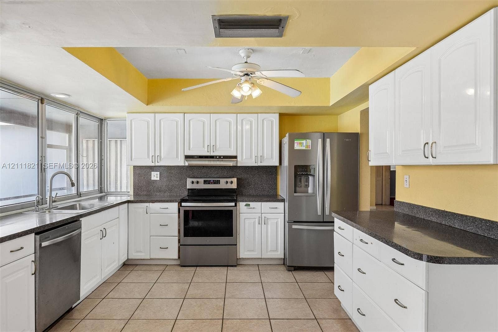 Kitchen featuring white cabinetry, dark stone-finish countertops, stainless steel appliances, ceramic tile flooring, and a tray ceiling with a multi-light ceiling fan