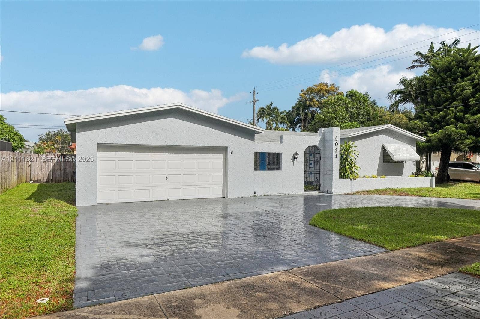 Single-story exterior featuring a light-toned stucco facade, dual-car garage, stamped concrete driveway, metal entryway gate, and awning-style window covers