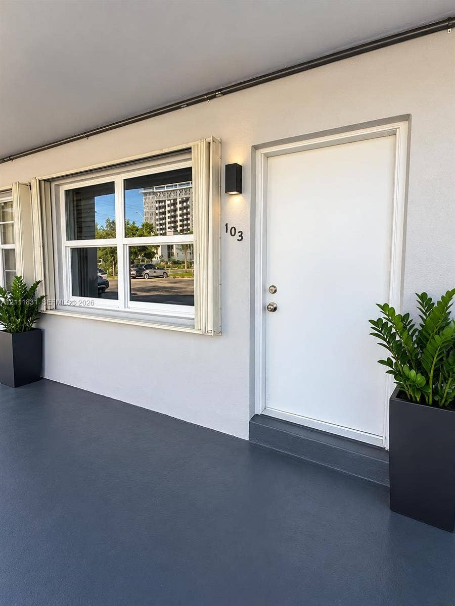 1629 N River Drive, #103, Miami, FL 33125 Exterior entryway featuring a white entry door, multi-pane window with retractable shutter, dark grey flooring, and a modern rectangular wall-mounted light fixture