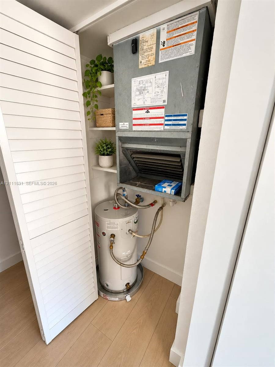 1629 N River Drive, #103, Miami, FL 33125 Utility closet featuring an electric water heater, wall-mounted air handling unit, and built-in shelving behind louvered doors with wood-finish flooring