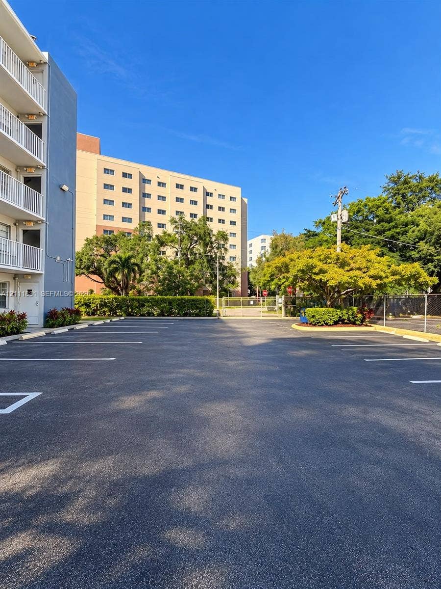 1629 N River Drive, #103, Miami, FL 33125 Paved surface parking area featuring marked spaces, perimeter metal fencing, and mature landscaping