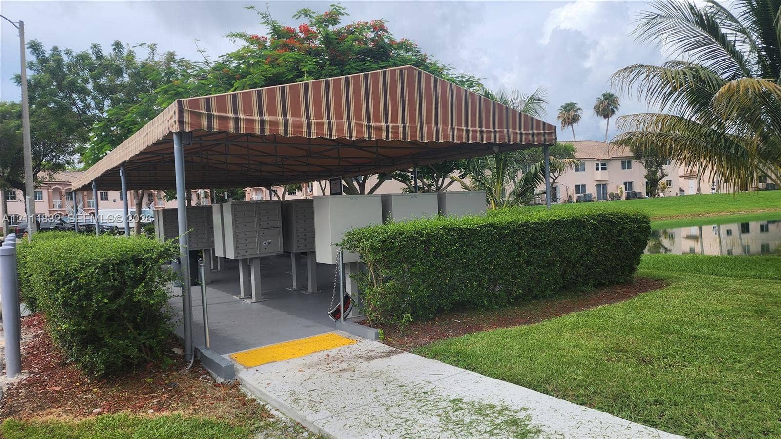 1250 SE 28th Court, #102, Homestead, FL 33035 Covered centralized mail delivery station with metal pedestal-mounted compartments, situated on a concrete pad with a striped fabric canopy