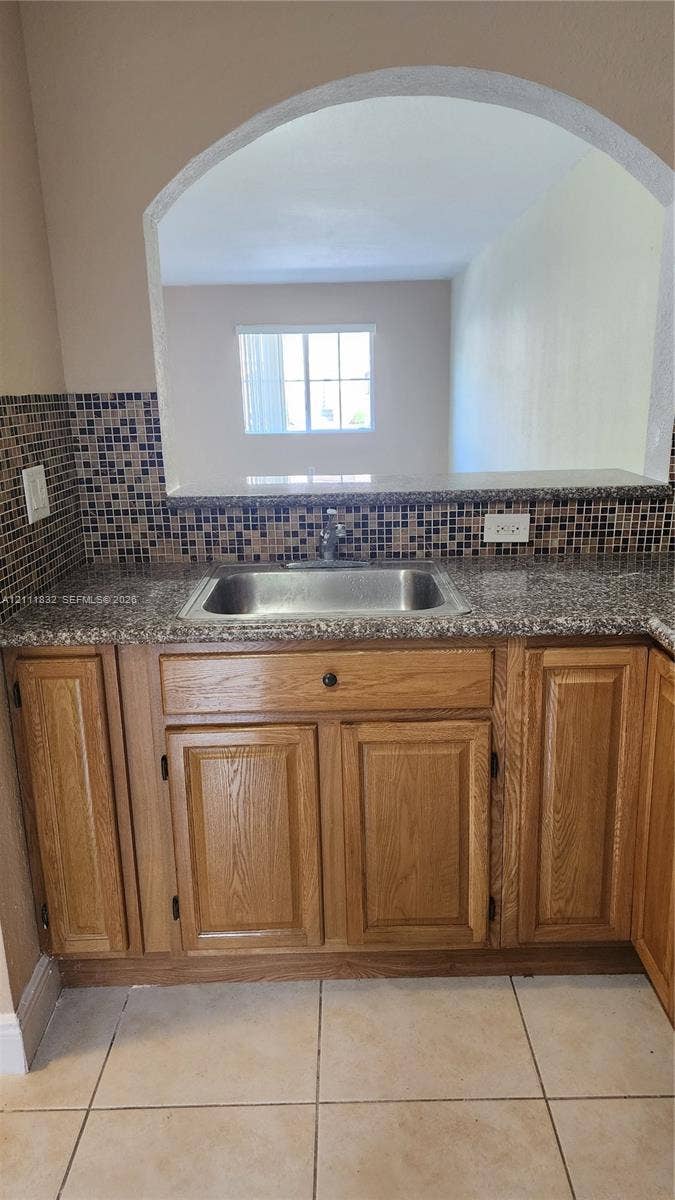 1250 SE 28th Court, #102, Homestead, FL 33035 Kitchen pass-through featuring an arched opening, stone-finish countertops, stainless steel sink, mosaic tile backsplash, and wood-finish cabinetry