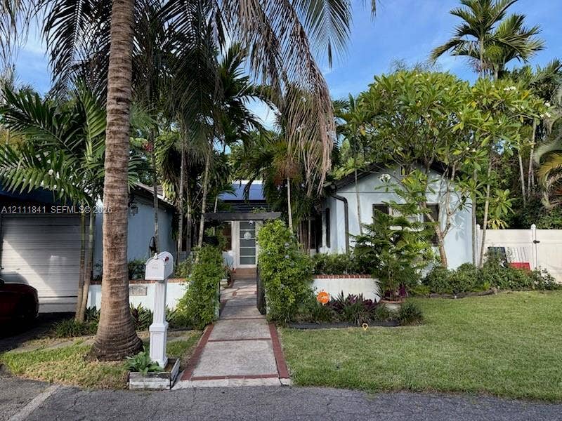 12015 NE 6th Avenue, Biscayne Park, FL 33161 Light-colored exterior siding with a covered entryway, paved walkway, brick-accented landscape border, and mature tropical landscaping