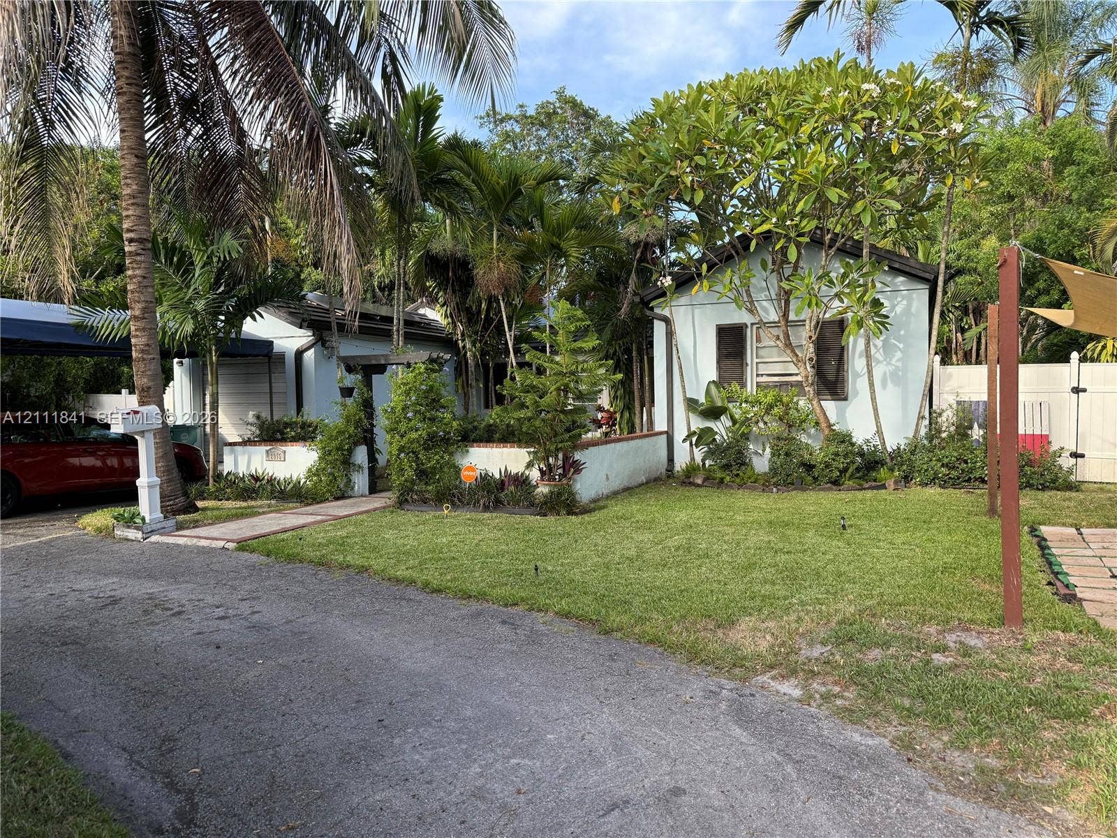 12015 NE 6th Avenue, Biscayne Park, FL 33161 Exterior facade featuring light-toned siding, a covered carport, masonry privacy wall, paved walkway, and mature landscaping