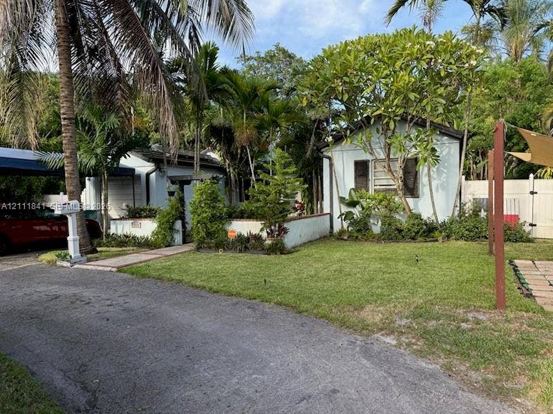 12015 NE 6th Avenue, Biscayne Park, FL 33161 Light-colored exterior siding with shuttered windows, complemented by a front yard with mature landscaping, a paved walkway, a metal-framed carport, and perimeter privacy fencing