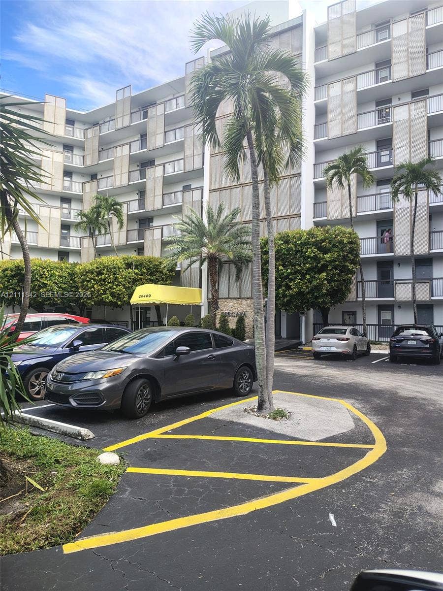 20400 W Country Club Drive, #303, Aventura, Aventura, FL 33180 Multi-story residential building featuring a concrete facade with decorative metal screening, private balconies, paved surface parking, and mature palm landscaping